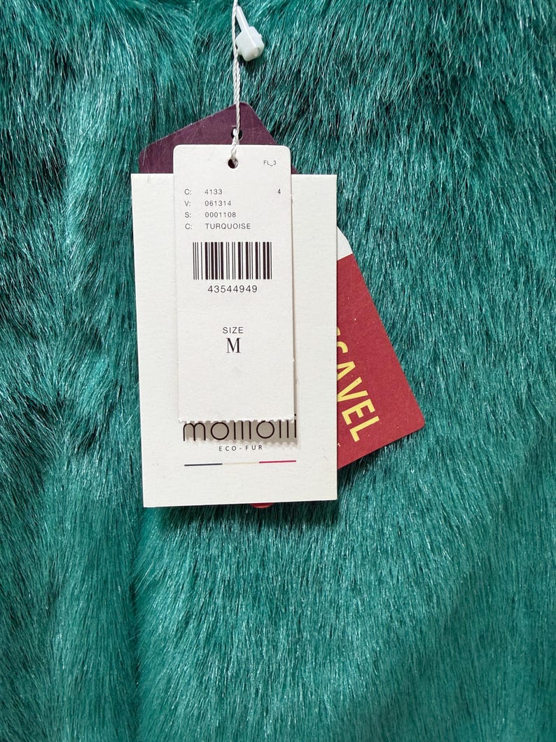 Op de afbeelding: Close-up van turquoise imitatiebontstof. Een wit label met zwarte tekst en een barcode is bevestigd, met de letter "M" voor de maat. Een rood label met het woord "CAVEL" is ook zichtbaar.