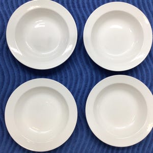 May include: A set of four white ceramic bowls arranged in a two-by-two grid. Each bowl features a wide, flat rim and a deep, circular basin. The surface of the bowls is smooth, glossy, and uniform in color, reflecting light on the interior curves.
