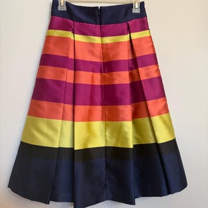 May include: A pleated skirt with horizontal stripes in shades of navy, black, yellow, orange, and purple. The skirt is hanging on a white hanger against a white background. The skirt has a navy blue waistband and a zipper closure.