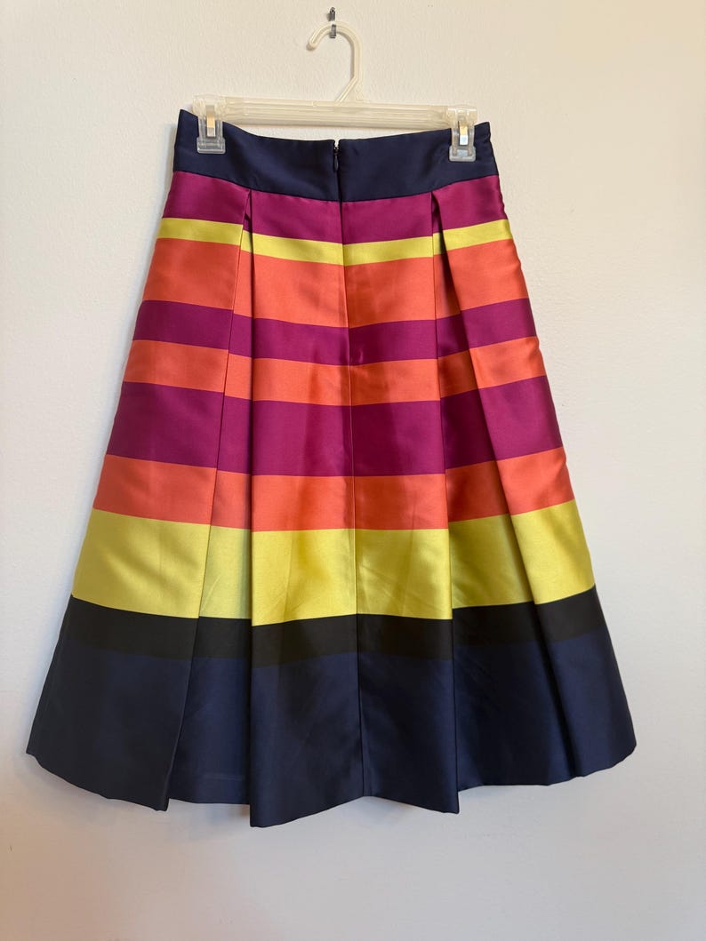 May include: A pleated skirt with horizontal stripes in shades of navy, black, yellow, orange, and purple. The skirt is hanging on a white hanger against a white background. The skirt has a navy blue waistband and a zipper closure.
