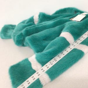 Op de afbeelding: Een turquoise imitatiebont kledingstuk met witte strepen. Het kledingstuk ligt op een wit oppervlak. Een liniaal is op het kledingstuk geplaatst en toont afmetingen in centimeters. Het bont ziet er zacht en pluche uit.