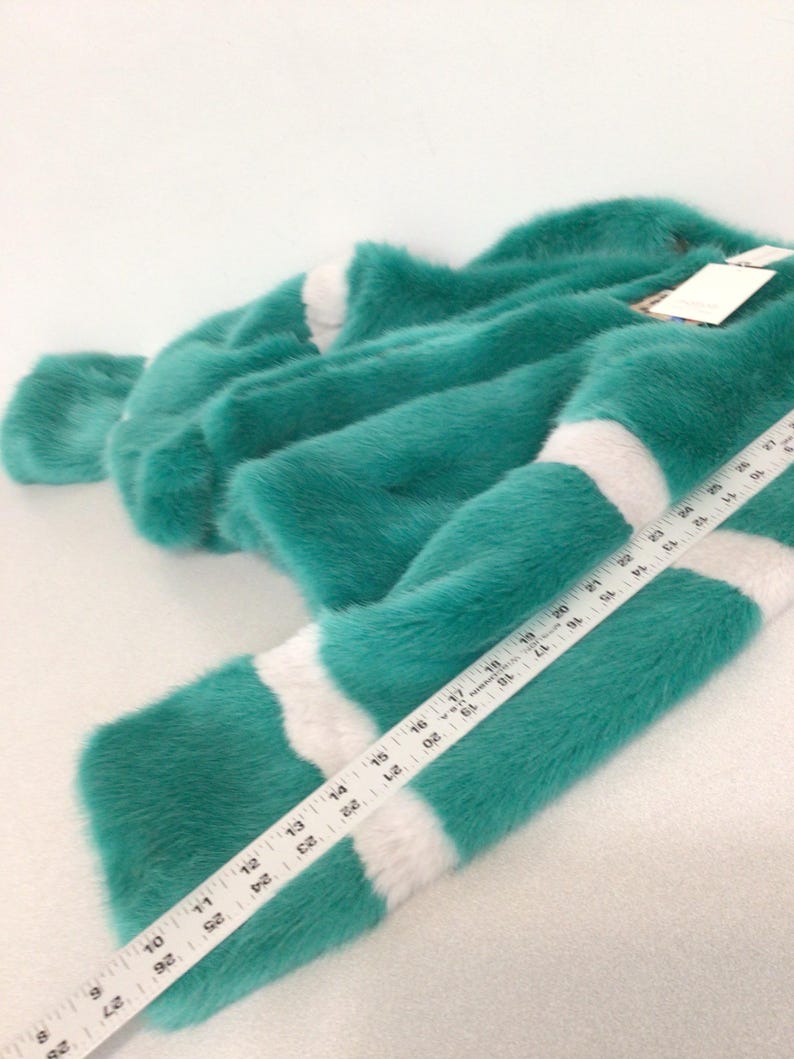 Op de afbeelding: Een turquoise imitatiebont kledingstuk met witte strepen. Het kledingstuk ligt op een wit oppervlak. Een liniaal is op het kledingstuk geplaatst en toont afmetingen in centimeters. Het bont ziet er zacht en pluche uit.