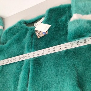 Op de afbeelding: Een teal faux fur jas met lange mouwen en een ronde halslijn. De mouwen hebben witte strepen bij de manchetten. Een liniaal toont de lengte van de jas. Een label is aan de jas bevestigd. De jas is ongeveer 51 cm lang.