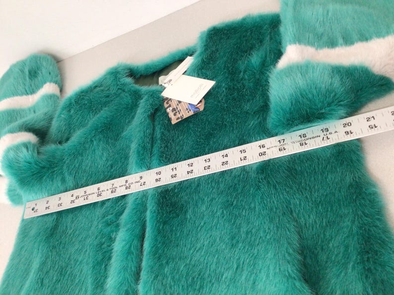 Op de afbeelding: Een teal faux fur jas met lange mouwen en een ronde halslijn. De mouwen hebben witte strepen bij de manchetten. Een liniaal toont de lengte van de jas. Een label is aan de jas bevestigd. De jas is ongeveer 51 cm lang.