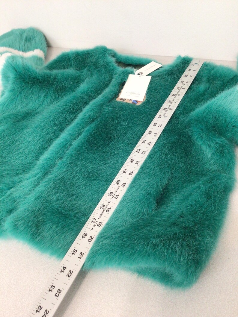 Op de afbeelding: Een teal-kleurige imitatiebontjas met een ronde hals en lange mouwen. Een liniaal ligt over de jas en meet ongeveer 61 cm. Een bijpassende teal-kleurige hoed met witte strepen is zichtbaar. Een label is bevestigd.