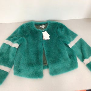Op de afbeelding: Een turquoise imitatiebontjas met een ronde halslijn en een open voorkant. De mouwen hebben twee witte strepen. Een label is bevestigd bij de halslijn. De jas is een opvallend kledingstuk.