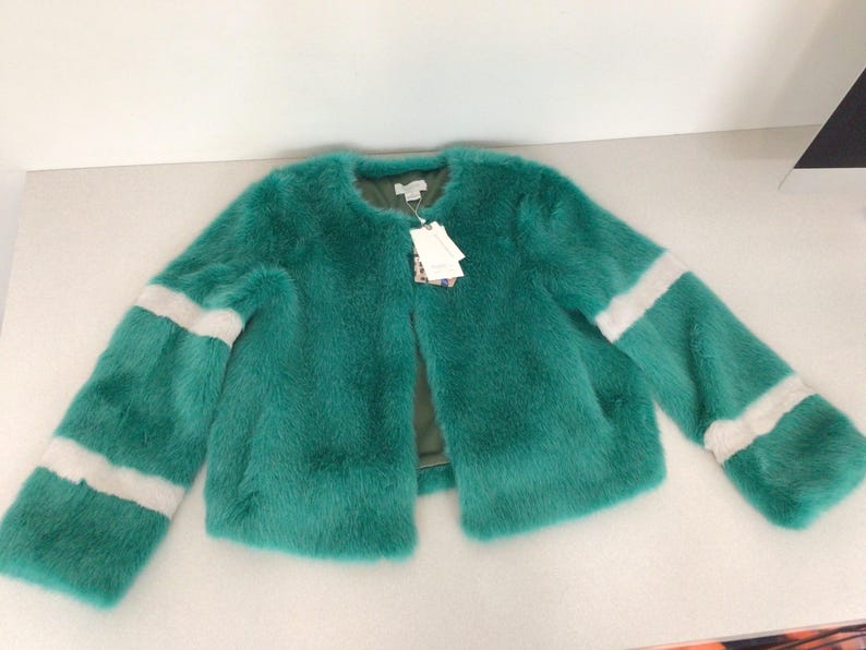 Op de afbeelding: Een turquoise imitatiebontjas met een ronde halslijn en een open voorkant. De mouwen hebben twee witte strepen. Een label is bevestigd bij de halslijn. De jas is een opvallend kledingstuk.