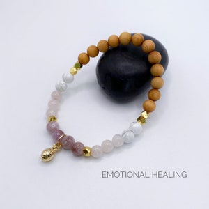 Könnte beinhalten: Ein Armband mit Holzperlen, weißen und rosafarbenen Steinen und einem goldenen Ananas-Anhänger. Das Armband liegt auf einem schwarzen Stein und der Text "EMOTIONAL HEALING" befindet sich unterhalb des Armbands.