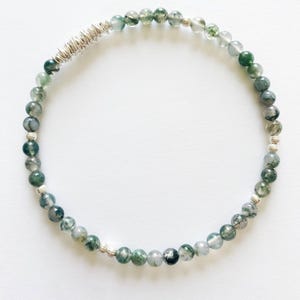 May include: A beaded bracelet featuring small, round, moss agate stones in shades of green and gray. The bracelet has silver-colored metal accents and a coiled design element. The bracelet is displayed on a white background.