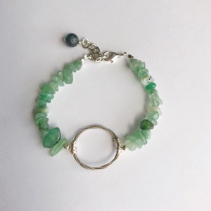 May include: A green gemstone bracelet with a silver circle charm and a silver clasp. The bracelet is made of small, irregular shaped green stones.