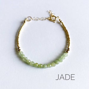 May include: A delicate gold bracelet with a row of light green jade beads. The bracelet has a gold clasp and is labeled "JADE".