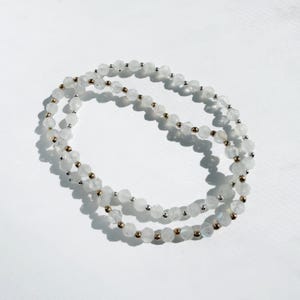 May include: A delicate necklace featuring small, faceted white beads interspersed with gold-colored beads. The necklace is draped on a white surface, creating a soft, elegant aesthetic. The beads are approximately 0.2 inches in diameter.