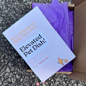 May include: A cardboard box containing a book titled "How to Repair or Make Your Own: Elevated Pet Dish!" with the artist's name, T. L. Ames Art. The book is on top of purple tissue paper.
