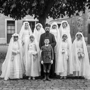 K&ouml;nnte beinhalten: Eine Gruppe von Kindern in wei&szlig;en Roben und Schleiern steht vor einem Mann in einer schwarzen Robe und einem Hut. Die Kinder halten kleine Blumenstr&auml;u&szlig;e. Die Gruppe steht vor einem Geb&auml;ude mit einer Steinfassade.
