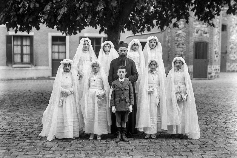 K&ouml;nnte beinhalten: Eine Gruppe von Kindern in wei&szlig;en Roben und Schleiern steht vor einem Mann in einer schwarzen Robe und einem Hut. Die Kinder halten kleine Blumenstr&auml;u&szlig;e. Die Gruppe steht vor einem Geb&auml;ude mit einer Steinfassade.