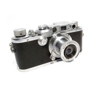 May include: A silver Leica camera with a black leather body and a silver lens. The lens has the text "Leitz Elmar f=5cm 1:3,5" and numbers around the lens barrel.