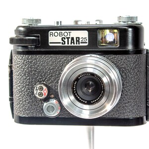 May include: A black Robot Star 25 camera with a silver lens. The camera has a textured black body and a silver Xenon 50/2.0 lens.