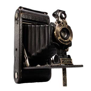 May include: A black vintage folding camera with a bellows and a lens with a gold-colored rim. The camera has a leather exterior and a brass nameplate that reads "No. 1A Autographic Kodak Jr."