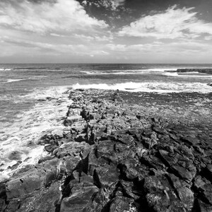 May include: A black and white photograph of a rocky coastline with waves crashing against the shore. The sky is cloudy and the water is choppy.
