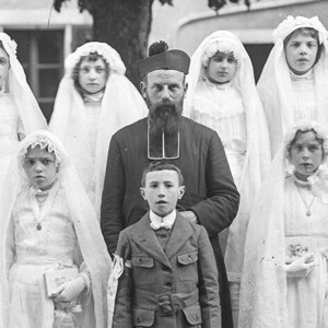 K&ouml;nnte beinhalten: Eine Gruppe von Kindern in wei&szlig;en Kleidern mit Schleiern steht vor einem Mann in einer schwarzen Robe und einem Hut. Die Kinder halten B&uuml;cher und l&auml;cheln. Der Mann hat einen Bart und schaut in die Kamera.