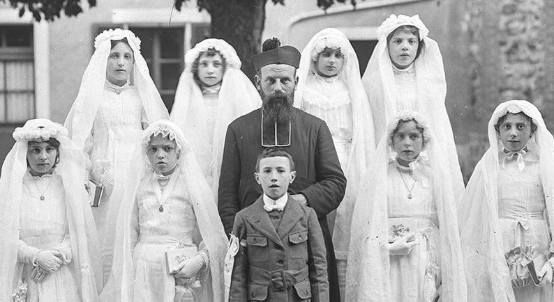 K&ouml;nnte beinhalten: Eine Gruppe von Kindern in wei&szlig;en Kleidern mit Schleiern steht vor einem Mann in einer schwarzen Robe und einem Hut. Die Kinder halten B&uuml;cher und l&auml;cheln. Der Mann hat einen Bart und schaut in die Kamera.