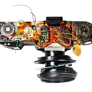 May include: A disassembled 35mm film camera with exposed wiring and circuitry. The camera is black and silver with a lens and a film advance lever. The camera is on a white background.