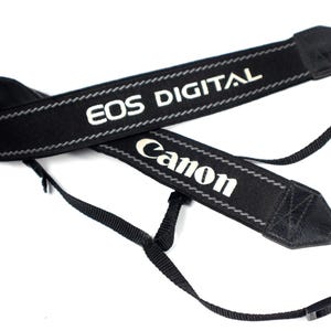 May include: Black camera strap with white text that reads "EOS DIGITAL" and "Canon".