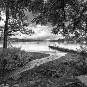 Op de afbeelding: Een zwart-witfoto van een meer met een houten steiger die het water in steekt. De steiger is gemaakt van verweerd hout en er missen een paar planken. Er zijn bomen aan de oever en een paar boten in de verte.