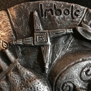 May include: A silver-toned metal plaque with a Celtic cross design and the word "Imbolc" inscribed on it. The cross is made of four arms with a square in the center.