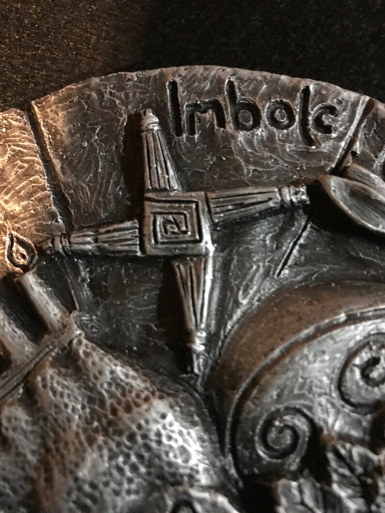 May include: A silver-toned metal plaque with a Celtic cross design and the word "Imbolc" inscribed on it. The cross is made of four arms with a square in the center.
