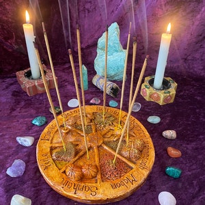 May include: A circular, tan incense holder with a star design and the names of eight sabbats. Incense sticks are placed in the holder, surrounded by colorful gemstones and two lit candles in decorative holders. The background is a purple velvet cloth.