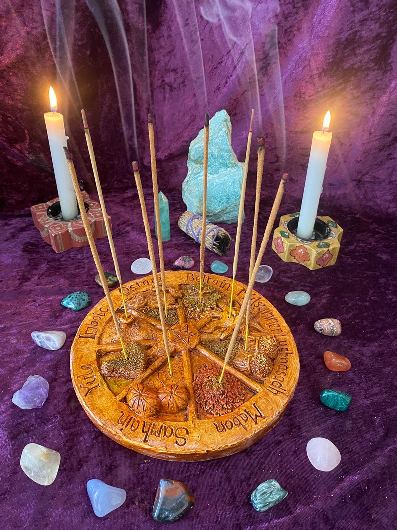 May include: A circular, tan incense holder with a star design and the names of eight sabbats. Incense sticks are placed in the holder, surrounded by colorful gemstones and two lit candles in decorative holders. The background is a purple velvet cloth.
