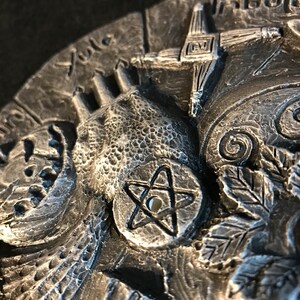 May include: A silver-toned metal disc with a pentagram design in the center. The disc is surrounded by various symbols and designs, including a hand, a cross, and a leaf.