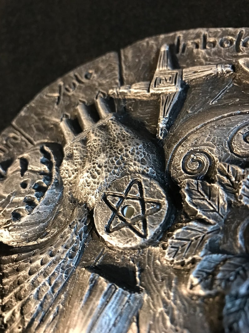 May include: A silver-toned metal disc with a pentagram design in the center. The disc is surrounded by various symbols and designs, including a hand, a cross, and a leaf.