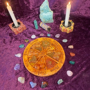 May include: A spiritual altar setup with a central wooden wheel featuring seasonal names, surrounded by crystals, candles in decorative holders, and a bundle of sage on a purple velvet surface. Two candles are lit.