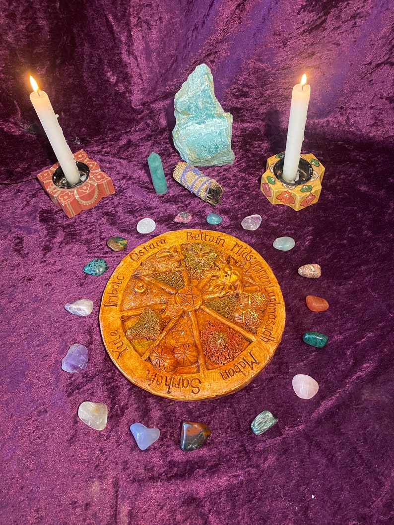 May include: A spiritual altar setup with a central wooden wheel featuring seasonal names, surrounded by crystals, candles in decorative holders, and a bundle of sage on a purple velvet surface. Two candles are lit.