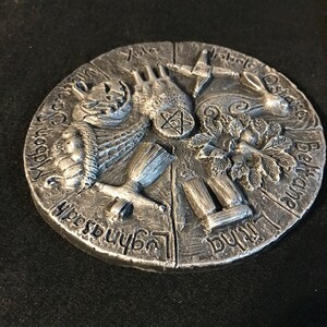 May include: A silver-colored circular metal plaque with raised relief carvings depicting the eight sabbats of the Wheel of the Year. The carvings are labeled with the names of the sabbats in a stylized font. The sabbats are Yule, Imbolc, Ostara, Beltane, Litha, Lammas, Mabon, and Samhain.