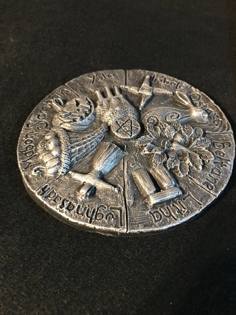May include: A silver-colored circular metal plaque with raised relief carvings depicting the eight sabbats of the Wheel of the Year. The carvings are labeled with the names of the sabbats in a stylized font. The sabbats are Yule, Imbolc, Ostara, Beltane, Litha, Lammas, Mabon, and Samhain.