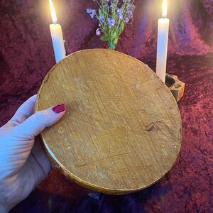May include: A round, wooden, gold-painted decorative tray with a textured surface. Two white candles are lit on either side of the tray, with a small bouquet of purple flowers in the background.