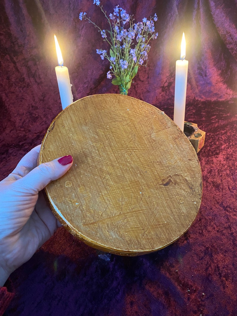 May include: A round, wooden, gold-painted decorative tray with a textured surface. Two white candles are lit on either side of the tray, with a small bouquet of purple flowers in the background.