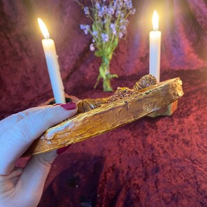 May include: A gold-colored, rectangular decorative tray with a textured surface. The tray is held in a hand with red nail polish. Two white candles are lit in the background, along with a small bouquet of purple flowers.