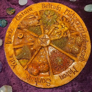 May include: A round, tan-colored decorative plaque with carved images and text. The plaque features a central pentagram and is divided into segments labeled with seasonal names like "Ostara" and "Beltane." Various gemstones surround the plaque on a purple surface.