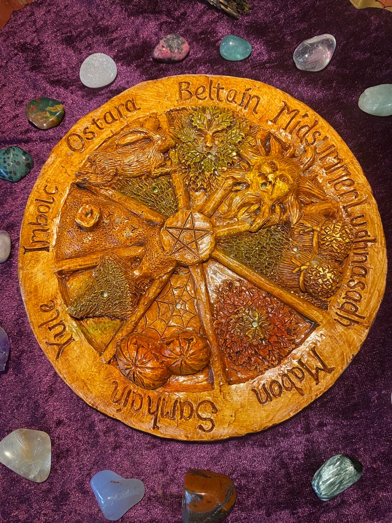 May include: A round, tan-colored decorative plaque with carved images and text. The plaque features a central pentagram and is divided into segments labeled with seasonal names like "Ostara" and "Beltane." Various gemstones surround the plaque on a purple surface.