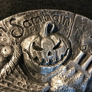 May include: A silver-toned metal ornament with a carved pumpkin face and the word "Samhain" inscribed around the top.