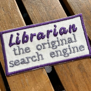 May include: Embroidered patch with the text "Librarian the original search engine" in purple and gray thread on a white background. The patch has a purple border and is rectangular in shape.