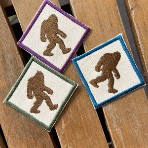 May include: Three embroidered patches featuring a brown silhouette of Bigfoot in different poses. The patches are square and have different colored borders: purple, blue, and green.