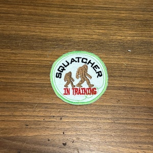 May include: Embroidered patch with a green border and the words "Squatcher In Training" in a circular design. The patch features two brown Bigfoot figures. The patch is round and has a white background.