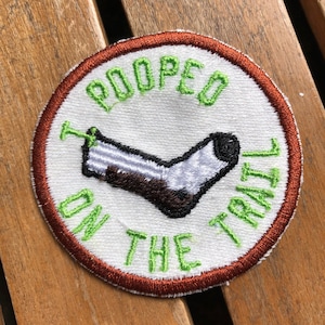 May include: A white embroidered patch with a brown and white sock design and the text "Pooped on the Trail" in green lettering. The patch has a brown border.