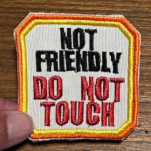 May include: Embroidered patch with the words "NOT FRIENDLY DO NOT TOUCH" in black and red lettering. The patch has a white background with vertical lines and is framed by yellow, orange, and red stitching.