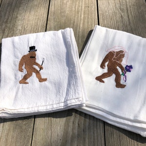 May include: Two white tea towels with embroidered images of Bigfoot. One Bigfoot is wearing a top hat and bowtie and holding a cane. The other Bigfoot is wearing a veil and holding a flower.